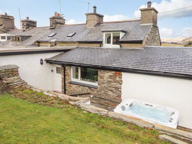 The exterior of a stone and white plaster house with a slate roof a grassy lawn and a hot tub outside at Ael Y Bryn in Ffestiniog