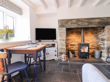 A room with a wooden dining table and chairs a stone fireplace with a wood burning stove a television on a white cabinet and a window with flowers on the sill at Ael Y Bryn in Ffestiniog