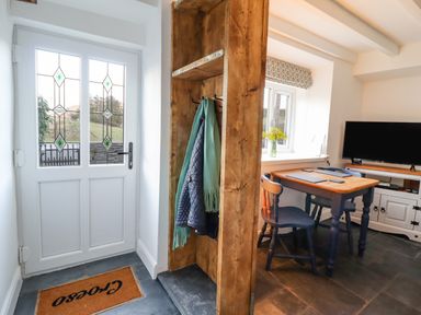 An entrance area with a glass paneled door a wooden coat rack next to a dining area with a table chairs window with flowers and a television screen at Ael Y Bryn in Ffestiniog
