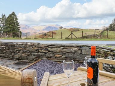 A wooden table with a bottle of wine and two wine glasses on a patio with a stone wall and a view of hills and trees at Ael Y Bryn in Ffestiniog
