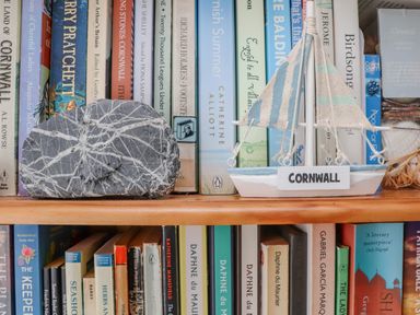 A bookshelf with books a decorative sailboat with Cornwall written on it and a large patterned rock