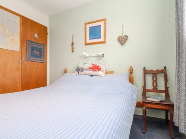 A bedroom with a double bed and striped bedding a wooden chair with books and a wooden wardrobe at Villa 29 in Camelford