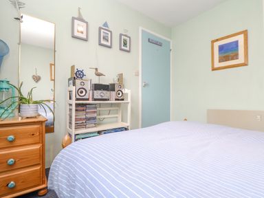 A bedroom with a bed with striped bedding a wooden chest of drawers a tall mirror a shelf with a stereo and cds and framed pictures on the wall at Villa 29 in Camelford