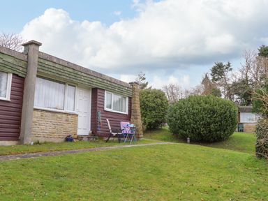 A single story house with a white door and windows and a garden with grass and large bushes at Villa 29 in Camelford