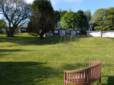A grassy outdoor area with a swing set wooden bench and single story houses at Villa 29 in Camelford