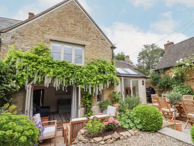 A stone house with green vines above an open door and an outdoor seating area with wooden chairs and plants at Lynton Cottage in Enstone