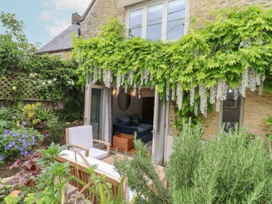 A garden seating area with white cushioned chairs and a wooden table outside a stone house with hanging white flowers at Lynton Cottage in Enstone