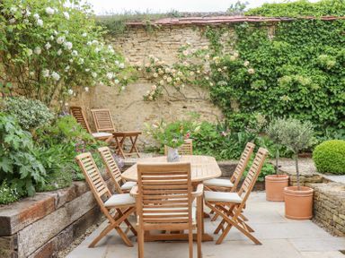 An outdoor patio with a wooden dining table and chairs surrounded by stone walls and plants at Lynton Cottage in Enstone