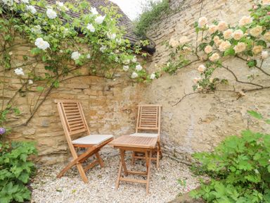 An outdoor seating area with two wooden chairs and a wooden table against stone walls with climbing plants and flowers at Lynton Cottage in Enstone