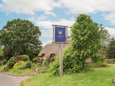 A blue sign for The Crown Inn on a wooden post near a thatched cottage and green trees in a rural setting at Lynton Cottage in Enstone