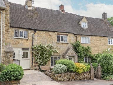 A stone cottage with a tiled roof and climbing plants on the walls surrounded by bushes and a small garden at Lynton Cottage in Enstone