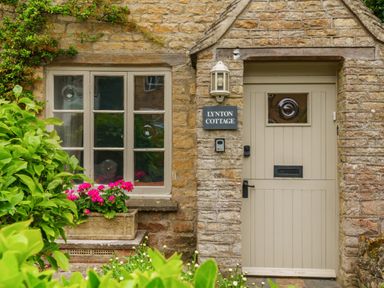 A stone cottage exterior with a beige door and window with flowers in front at Lynton Cottage Enstone