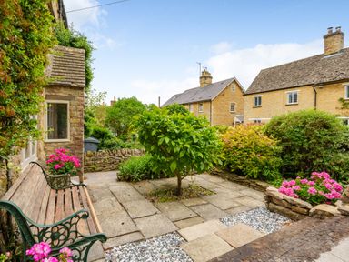A garden area with stone paving a bench small tree flowering plants and stone houses in the background at Lynton Cottage in Enstone