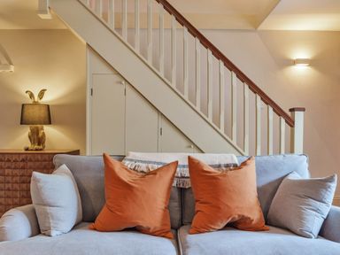 A sofa with orange and light grey cushions in front of a staircase with a wooden handrail at Lynton Cottage in Enstone