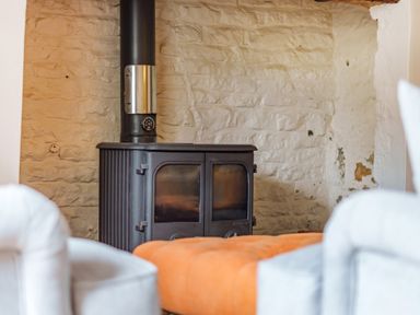 A black wood stove against a stone wall with white chairs and an orange cushion in front at Lynton Cottage in Enstone