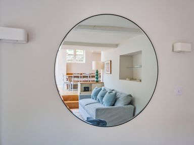 A round mirror reflecting a living room with a sofa and cushions and a dining table with chairs at Lynton Cottage in Enstone