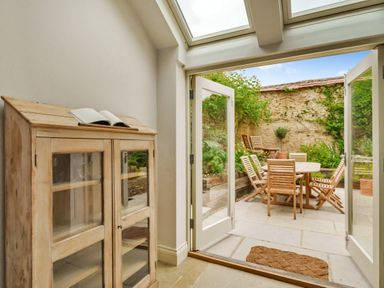 A room with a wooden cabinet and an open book next to open glass doors leading to a patio with wooden table and chairs at Lynton Cottage in Enstone