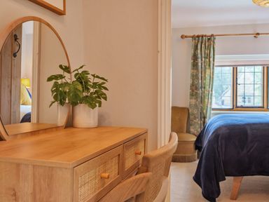 A wooden dressing table with a round mirror and a plant next to a chair and a bedroom with a bed and window at Lynton Cottage in Enstone
