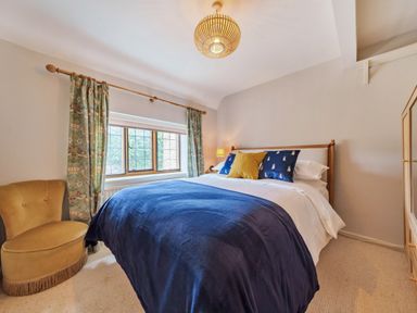 A bedroom with a bed covered in blue bedding two blue and one yellow pillows a yellow chair floral curtains and a wooden wardrobe at Lynton Cottage in Enstone