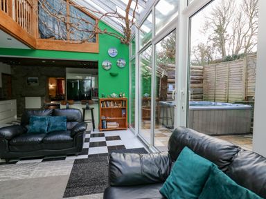 A sunroom with black leather sofas and green pillows next to glass doors and a hot tub outside at Cringoed House in Llanarth near Aberaeron