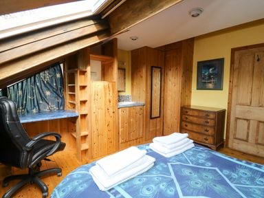 A bedroom with a bed covered in blue peacock patterned bedding and folded towels on top wooden wardrobe a desk with a black office chair and a window with blue patterned curtains at Cringoed House in Llanarth near Aberaeron