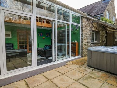 A glass door leads to a living area with black leather chairs and green walls next to a stone building with a hot tub outside at Cringoed House in Llanarth near Aberaeron