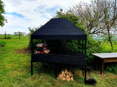 An outdoor black metal barbecue grill with bags of charcoal and wood logs stacked underneath in a grassy area at Cringoed House Llanarth near Aberaeron