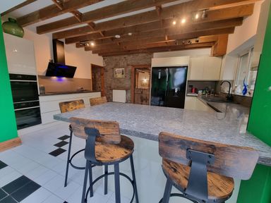 A kitchen with a stone wall and wooden beams a large island with wooden stools a black refrigerator and white cabinets at Cringoed House Llanarth near Aberaeron