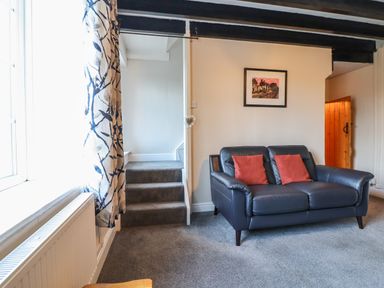 A room with a blue leather sofa with two red cushions a framed picture on the wall stairs with carpet and a window with patterned curtains at Ynys in Dyffryn Ardudwy