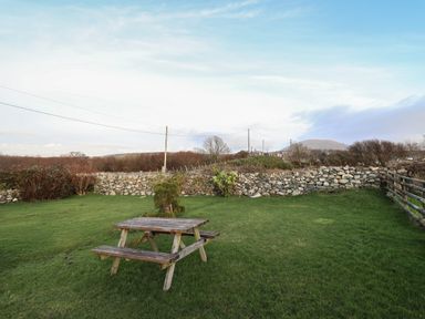 An outdoor grassy area with a wooden picnic table and bench surrounded by a stone wall and wooden fence at Ynys in Dyffryn Ardudwy