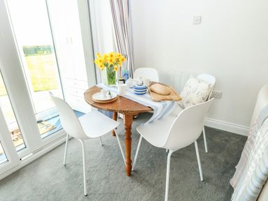 A dining area with a round wooden table set with dishware yellow flowers and bread surrounded by white chairs and a bench at Gull Cottage in Kessingland