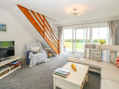 A living room with a beige sofa and pillows a coffee table with books and lemons a striped covered armchair a television a wooden staircase and sliding glass doors at Gull Cottage in Kessingland