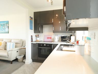 A kitchen with black cabinets, a stove, a sink, and a white countertop next to a living room with a beige sofa and a painting on the wall at Gull Cottage in Kessingland