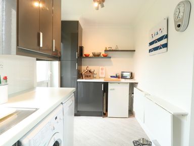 A kitchen with grey cabinets white countertops a washing machine a small refrigerator a toaster shelves with bowls and jars a wall clock and a wall hanging at Gull Cottage in Kessingland