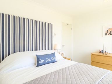 A bedroom with a striped headboard a bed with a pillow that says Sun Sea Sleep a wooden dresser and a door at Gull Cottage in Kessingland
