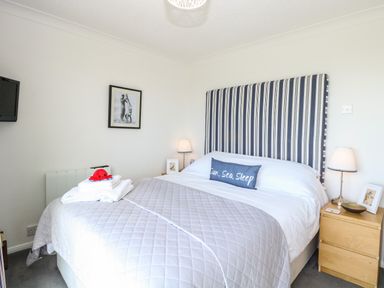 A bedroom with a bed featuring a striped headboard bedside tables with lamps and framed pictures at Gull Cottage in Kessingland