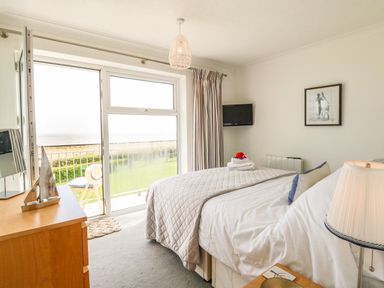A bedroom with a bed a dresser and a door opening to a balcony with a view of grass and sea at Gull Cottage in Kessingland