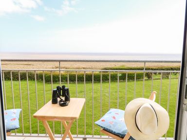 A balcony with a wooden table and chairs overlooking a grassy area a beach and the sea at Gull Cottage in Kessingland
