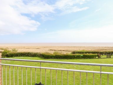 A view of a beachfront with a grass lawn and hedges from behind a metal railing at Gull Cottage in Kessingland