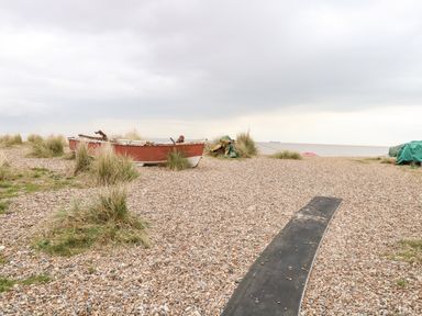 A pebble beach area with a red and white boat surrounded by grass and a path made of a black mat near the sea at Gull Cottage in Kessingland