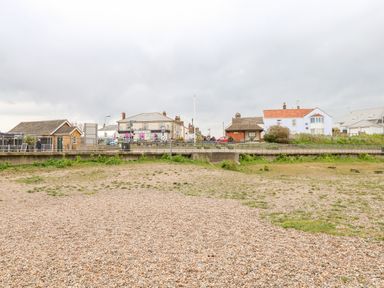 A pebble beach with grassy patches and houses along a road in the background at Gull Cottage in Kessingland