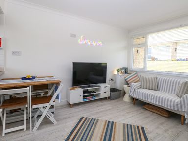 A living room with a TV on a white stand a wooden table with chairs and a striped sofa at Dollys Lookout in Kessingland