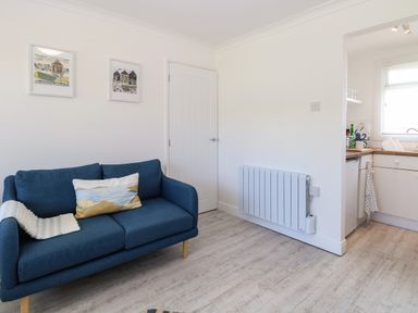 A small living room with a blue sofa and framed pictures on white walls next to a kitchen at Dollys Lookout in Kessingland