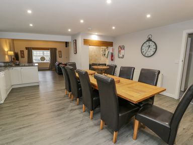 A dining area with a long wooden table and eight black chairs near a kitchen and living room at Bodrual Cottage in Rhosbodrual near Caernarfon