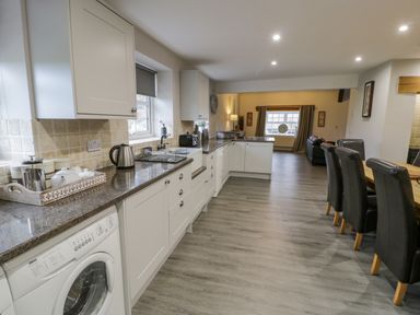 A kitchen with white cabinets and appliances including a washing machine and kettle and a dining area with black chairs at Bodrual Cottage in Rhosbodrual near Caernarfon