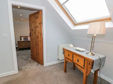 A hallway with a wooden side table holding a lamp and decorative items next to a skylight window and an open door showing a bedroom with a wooden bed frame at Bodrual Cottage in Rhosbodrual near Caernarfon