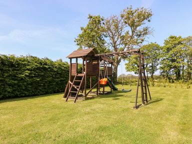 A wooden playset with swings and a slide on grass surrounded by trees and hedges at Bodrual Cottage in Rhosbodrual near Caernarfon