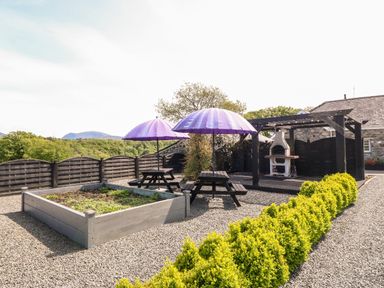 An outdoor seating area with two picnic tables with purple umbrellas a raised garden bed and a pergola with a barbecue at Bodrual Cottage in Rhosbodrual near Caernarfon