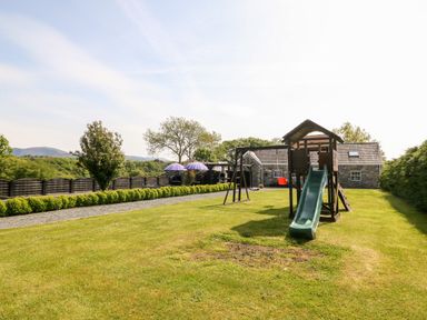 A grassy backyard with a playground set featuring a slide and swings near a stone house at Bodrual Cottage in Rhosbodrual near Caernarfon
