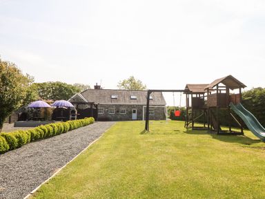 A garden with a playset slide and swings a gravel path with shrubs and a stone house in the background at Bodrual Cottage in Rhosbodrual near Caernarfon